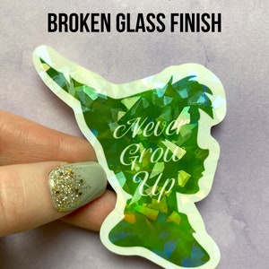 Pu&ograve; includere: Un adesivo olografico con finitura vetro rotto, raffigurante una silhouette verde di un personaggio con la scritta "Never Grow Up". L'adesivo &egrave; tenuto su uno sfondo viola chiaro. Il testo "EXAMPLE OF BROKEN GLASS FINISH" &egrave; in alto.