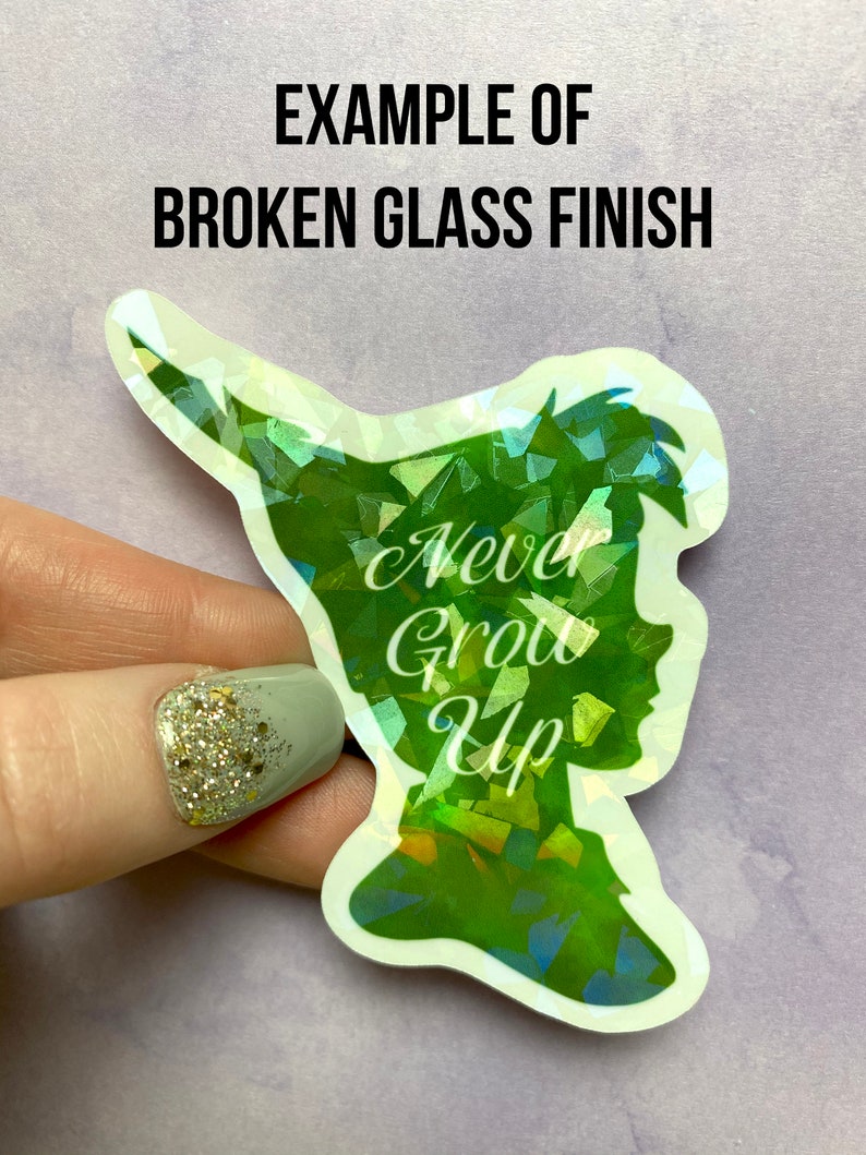 Pu&ograve; includere: Un adesivo olografico con finitura vetro rotto, raffigurante una silhouette verde di un personaggio con la scritta "Never Grow Up". L'adesivo &egrave; tenuto su uno sfondo viola chiaro. Il testo "EXAMPLE OF BROKEN GLASS FINISH" &egrave; in alto.