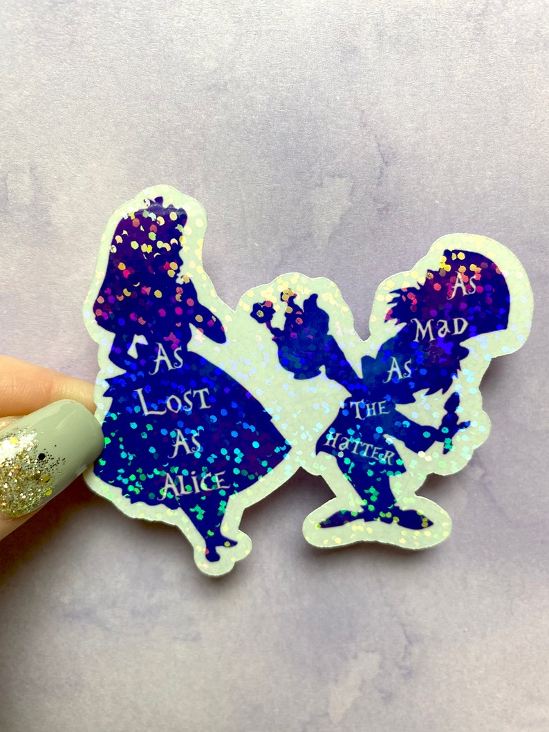 Puede incluir: Un par de pegatinas de purpurina iridiscente con siluetas de Alicia y el Sombrerero Loco de Alicia en el Pa&iacute;s de las Maravillas. El texto en las pegatinas dice "As Lost As Alice" y "As Mad As The Hatter".