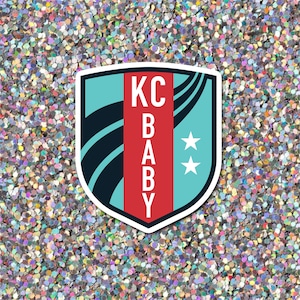 May include: A blue and red shield-shaped sticker with the text "KC BABY" and two white stars. The sticker is on a silver glitter background.