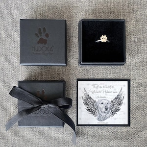 May include: A black box with a white paw print logo that says "HUDOCA Human-Dog-Cat" and a black ribbon. A smaller black box with a silver ring with a gold paw print charm. A card with a black and white illustration of a dog with wings and the text "Thank you so much from Angel who is Hudoca's muse in heaven... www.hudocan.com"