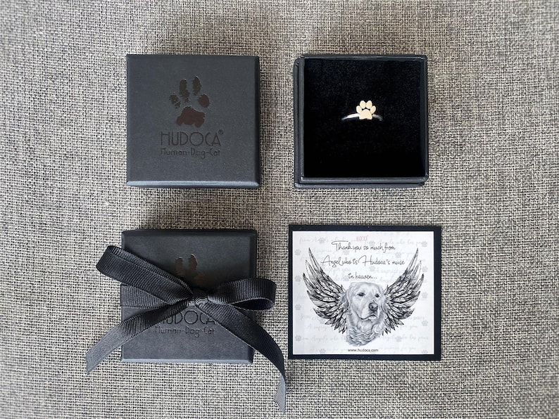 May include: A black box with a white paw print logo that says "HUDOCA Human-Dog-Cat" and a black ribbon. A smaller black box with a silver ring with a gold paw print charm. A card with a black and white illustration of a dog with wings and the text "Thank you so much from Angel who is Hudoca's muse in heaven... www.hudocan.com"