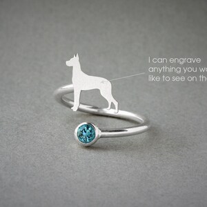 May include: A silver-toned bypass ring featuring a silhouette of a dog and a round, faceted blue gemstone. The ring includes text that reads: I can engrave anything you would like to see on the ring, with a line pointing to the dog silhouette.