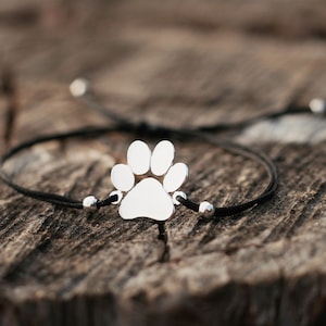 May include: A black string bracelet with a silver paw print charm. The bracelet is tied in a knot and has silver beads on either side of the charm.