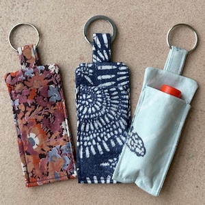May include: Three fabric keychain holders with silver key rings. One has a floral pattern in shades of red, orange, and purple. Another has a navy blue and white spiral design. The third is light blue with a butterfly print and holds a red lip balm.