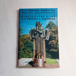 Op de afbeelding: Boekomslag met een stenen sculptuur van een man tegen een blauwe lucht en bomen. De titeltekst is in meerdere talen, waaronder het Nederlands. De sculptuur staat in een park met een pad en bloemen.