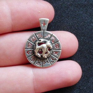 May include: Silver pendant featuring a zodiac wheel with the Leo symbol in the center. The wheel has all 12 zodiac signs around the edge.