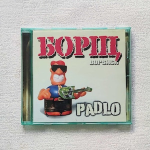 Op de afbeelding: Een cd-hoesje met een stripfiguur met een zonnebril en een gitaar. De cd-hoes toont de naam "BORII" in rode letters, met "PADLO" eronder. De hoes heeft een transparante, lichtgroene kleur.