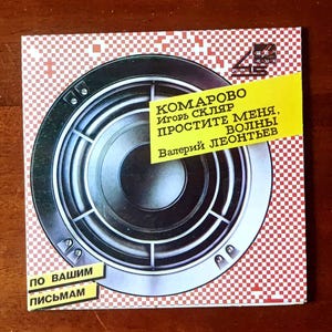 Peut inclure: Pochette d'album carrée avec un motif circulaire argenté et noir, un rectangle jaune avec du texte russe et un fond à carreaux rouges et blancs. Le texte sur le rectangle jaune se lit "KOMAROVO, IGOR SKLYAR, PROSTITE MENYA, VOLNY, VALERY LEONTIEV."