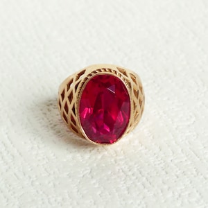 Op de afbeelding: Een gouden ring met een grote, ovale, rode edelsteen gezet in een decoratief, opengewerkt ontwerp.