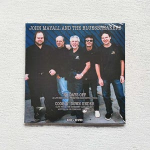May include: CD + DVD of John Mayall and The Bluesbreakers. The album cover features a group photo of the band. Text on the cover reads "NO DAYS OFF" and "COOKIN' DOWN UNDER" (Live from the Basement Club Sydney, Australia on February 12th, 2003).