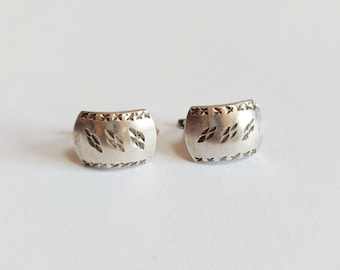 May include: A pair of silver-toned cufflinks. Each cufflink has a curved rectangular shape with a decorative pattern of leaf-like shapes and small cross-like designs. The cufflinks are set against a white background.