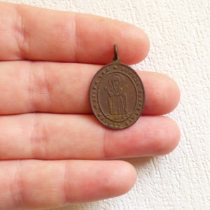 Puede incluir: Un peque&ntilde;o colgante de forma ovalada de color bronce con una imagen de un santo y texto cir&iacute;lico. El colgante tiene un bucle en la parte superior para colgar.