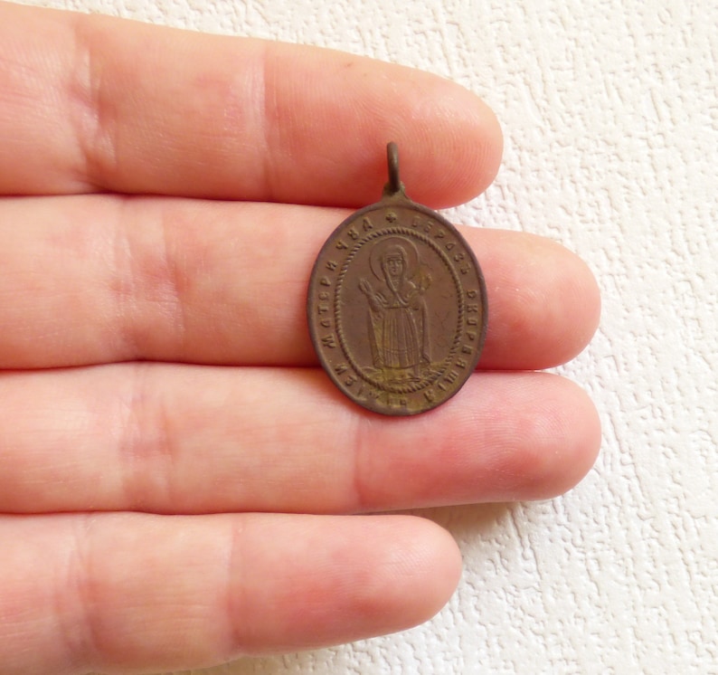 Puede incluir: Un peque&ntilde;o colgante de forma ovalada de color bronce con una imagen de un santo y texto cir&iacute;lico. El colgante tiene un bucle en la parte superior para colgar.