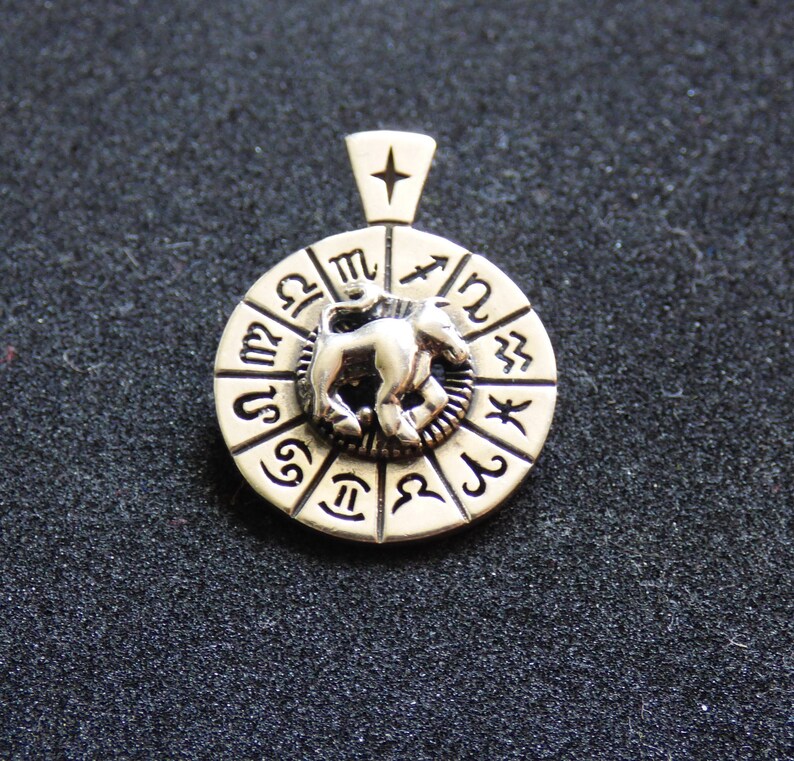 May include: Silver pendant featuring a circular zodiac wheel with a silver bull in the center. The wheel is decorated with the symbols of the twelve zodiac signs.