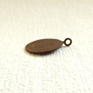 Puede incluir: Una peque&ntilde;a medalla ovalada de color bronce con un dise&ntilde;o en relieve y un bucle para colgar. La medalla tiene un aspecto desgastado y envejecido.