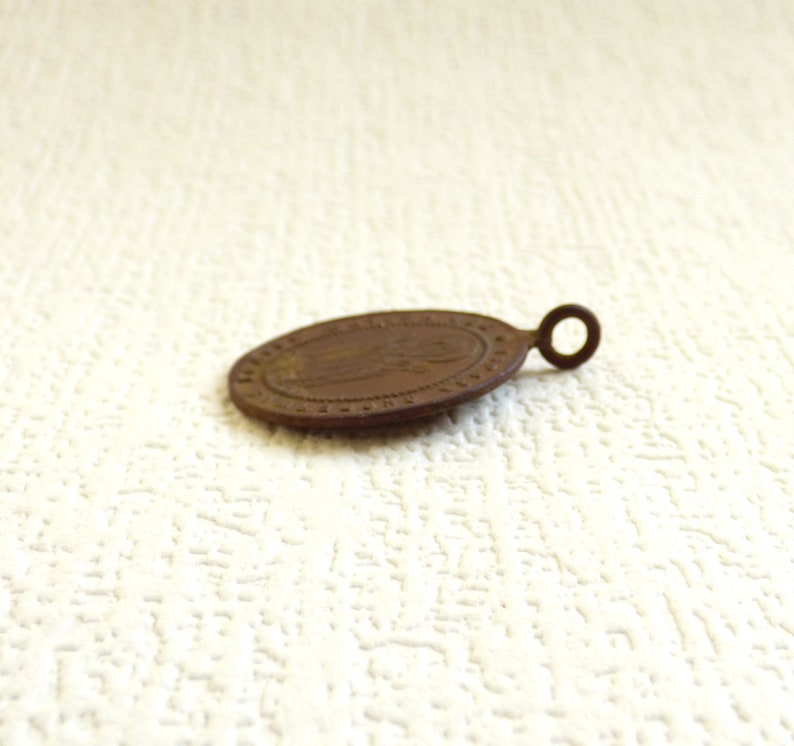 Puede incluir: Una peque&ntilde;a medalla ovalada de color bronce con un dise&ntilde;o en relieve y un bucle para colgar. La medalla tiene un aspecto desgastado y envejecido.