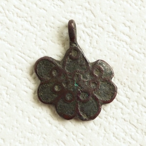 May include: A bronze pendant in the shape of a flower with a loop at the top for hanging. The flower has a circular design with a central circle and smaller circles around it.