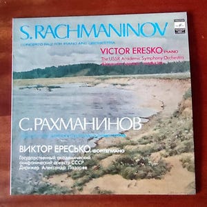 Puede incluir: Una portada de álbum de vinilo vintage con el nombre "S. Rachmaninov" en azul. La portada incluye texto en inglés y ruso, con una imagen de paisaje de un lago y la costa. El álbum presenta a Victor Eresko al piano.