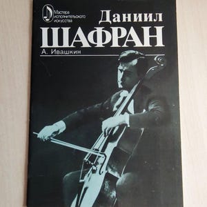 Op de afbeelding: Zwart-wit boekcover met een man die cello speelt. De titel "Даниил Шафран" staat in grote witte Cyrillische letters. De cover toont ook de tekst "Мастера исполнительского искусства" en de naam van de auteur.