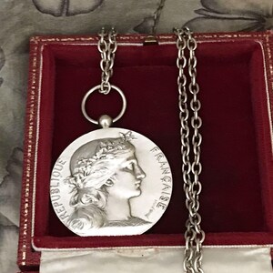May include: A silver-colored pendant necklace with a chain, displayed in a red box. The pendant features a profile of a woman with a laurel wreath and the words "REPUBLIQUE FRANCAISE". The necklace is on a patterned background.