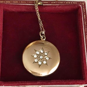 May include: A gold locket necklace with a starburst design. The round pendant has a starburst pattern with clear, sparkling stones. The necklace is presented in a red velvet jewellery box, highlighting its classic appeal.