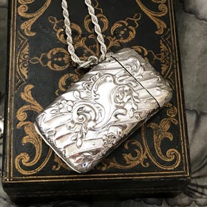 May include: A rectangular silver pendant with an ornate floral design and a silver chain. The pendant is highly detailed with engraved patterns and is resting on a dark, patterned surface.