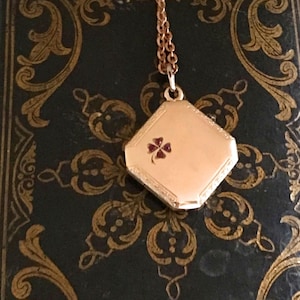May include: A gold-toned octagonal locket pendant suspended from a matching link chain. The locket features a small, dark red four-leaf clover inlay on its front surface, with delicate engraved detailing along the beveled edges of the frame.