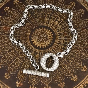 May include: A silver bracelet with a toggle clasp. The bracelet has a chain link design. The toggle clasp features a round, crystal-studded loop and a crystal-studded bar. The bracelet is displayed on a brown, ornate surface.