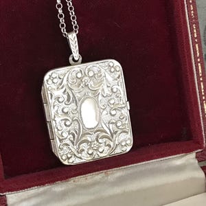 May include: A rectangular silver locket featuring an intricate embossed floral and scrollwork pattern with a smooth central oval panel. The locket is suspended from a silver chain and is presented inside a deep red velvet-lined jewelry box.