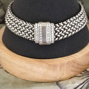 May include: A silver-toned woven metal bracelet featuring a rectangular center clasp. The clasp is accented with two vertical rows of small, clear, sparkling stones flanking a smooth, polished silver-toned rectangular center panel.