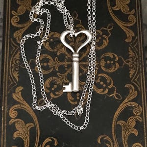 May include: A silver-colored key pendant necklace with a heart-shaped top. The key hangs on a delicate chain, resting on a dark, patterned surface. The necklace is a romantic accessory.