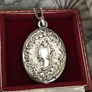 May include: Oval silver pendant necklace with ornate floral and scrollwork detailing. The pendant is suspended from a delicate chain and rests inside a red velvet jewelry box. The pendant has a polished, reflective surface.