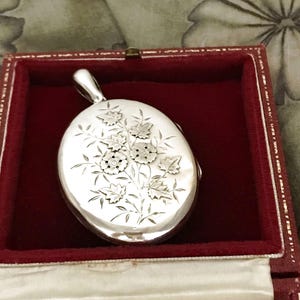 May include: Oval silver locket with floral engraving, displayed in a red jewelry box. The locket features a detailed design of flowers and leaves. The box has a white satin interior.