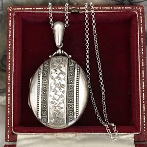 May include: An oval silver locket pendant featuring a central panel with an engraved floral pattern flanked by two vertical bands of beaded detailing. The locket is suspended from a silver rolo chain and rests inside a square red velvet-lined box.