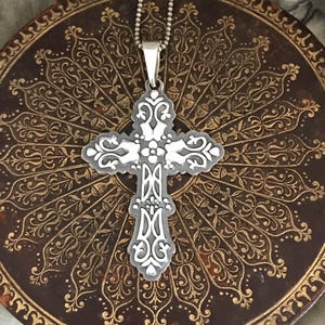 May include: Ornate silver-tone cross pendant with white accents, suspended from a delicate chain. The cross features intricate filigree detailing and a central floral design. The pendant rests on a dark brown, patterned surface.