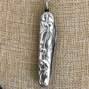 May include: A silver pendant necklace with a decorative pocket knife design. The knife has detailed floral patterns and hangs from a delicate silver chain. The pendant is displayed against a neutral, textured background.