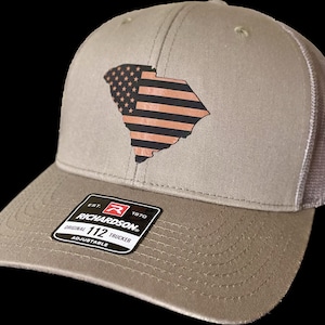 Puede incluir: Una gorra de camionero de color beige y gris con un diseño de bandera estadounidense en blanco y negro del estado de Carolina del Sur. La gorra tiene una correa ajustable y una etiqueta Richardson 112.