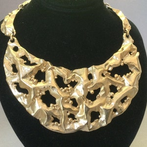 May include: A gold-toned statement necklace with a textured, abstract design. The necklace is made of metal and features small, round beads throughout the design.