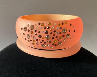 May include: A peach-colored bangle bracelet with a scattered pattern of clear and brown rhinestones.