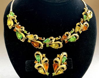May include: A gold-toned necklace and matching earrings set. The jewelry features a leaf-like design with teardrop-shaped gemstones in shades of green, yellow, and orange. The set is displayed on a black velvet background.