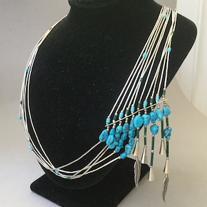 May include: A silver necklace with multiple strands, adorned with turquoise stones and green beads. The necklace features dangling turquoise clusters, silver cones, and feather charms, displayed on a black mannequin.
