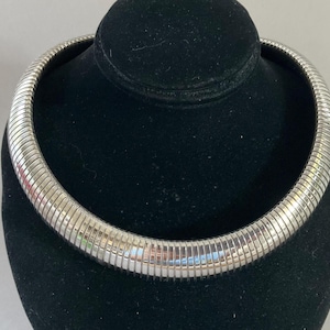 May include: A silver-colored, ribbed collar necklace displayed on a black velvet bust. The necklace has a flexible, tubular design with a polished finish. The necklace is a choker style, designed to sit close to the neck.