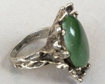 May include: A silver ring with a green stone set in a detailed, organic design. The band is marked "STERLING".