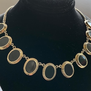 May include: A gold-tone chain necklace featuring nine oval-shaped black cabochon stones set in gold-tone metal bezels. The necklace is arranged in a graduated pattern with the largest oval stones centered and smaller stones along the sides.