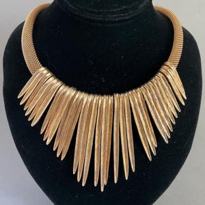 May include: Gold-toned statement necklace. The necklace features a mesh chain and a series of vertical, elongated, pointed gold-colored elements that fan out from the center. The necklace is displayed on a black mannequin bust.