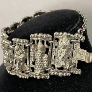 May include: Silver-toned bracelet featuring detailed rectangular links with various embossed designs. The bracelet includes images of a person, a tower, and a face. The clasp is a simple hook and eye closure. The bracelet is displayed on a black surface.