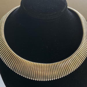May include: A silver-toned collar necklace with a ribbed design. The necklace is displayed on a black velvet bust. The ribbed design creates a textured, eye-catching appearance. The necklace is a statement piece.