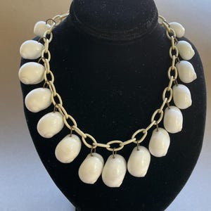 May include: A necklace featuring a series of smooth, white, oval-shaped shells suspended from a cream-colored plastic chain. Each shell is attached to the chain with a small, thin, dark metal jump ring, creating a uniform, repeating pattern.