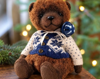 May include: A brown plush teddy bear wearing a cream and blue knit cardigan with a snowflake pattern. The bear has a dark brown nose and eyes, and a blue jingle bell tied around its neck with a thin cream ribbon, sitting in a seated position.